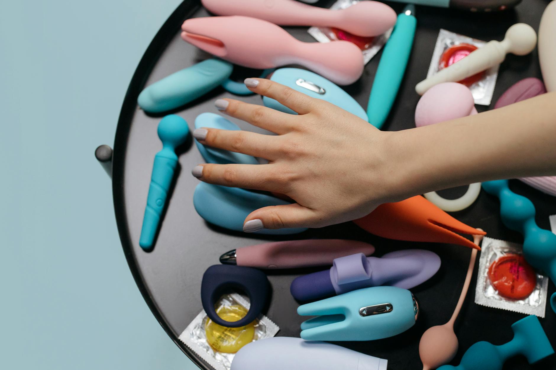 Duas mãos segurando vibradores de silicone rosa e azul contra fundo pastel