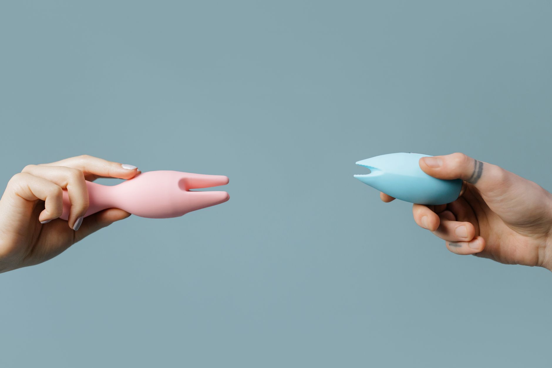 Duas mãos segurando vibradores de silicone rosa e azul contra fundo pastel